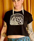 Person wearing a black As Above, So Below Crop Top (White) with rolled sleeves and graphic design, paired with striped blue and black pants, standing against a gold curtain backdrop.
