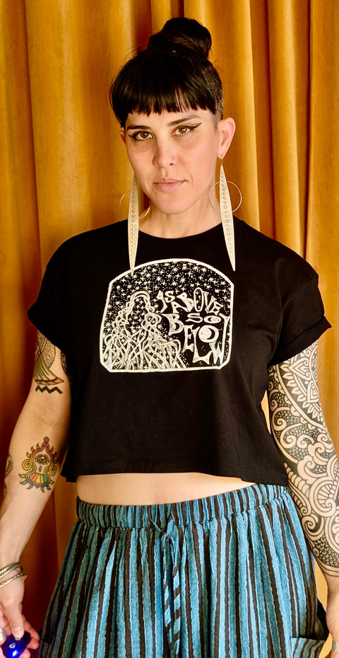 Person wearing a black As Above, So Below Crop Top (White) with rolled sleeves and graphic design, paired with striped blue and black pants, standing against a gold curtain backdrop.