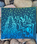 As Above, So Below — Ombre Linocut Print featuring hand-pulled black ink design of a mystical figure surrounded by stars on hand-painted blue-green watercolor paper, set on natural stones.