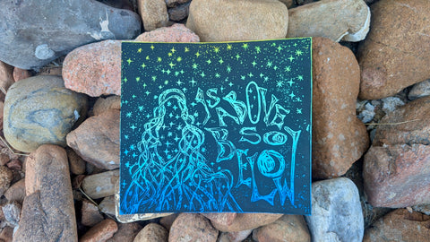 As Above, So Below — Ombre Linocut Print featuring hand-pulled black ink design of a mystical figure surrounded by stars on hand-painted blue-green watercolor paper, set on natural stones.
