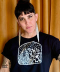 Woman with dark hair and winged eyeliner wearing large pointed silver earrings and an "As Above, So Below Crop Top (White)" black crop top with white celestial design against a mustard curtain background