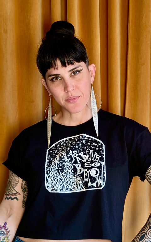 Woman with dark hair and winged eyeliner wearing large pointed silver earrings and an "As Above, So Below Crop Top (White)" black crop top with white celestial design against a mustard curtain background