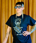 Model wearing a black "Be a Lilith Tee" with a bold owl and tentacle design, oversized glasses, large earrings, and tattooed arms against a mustard yellow curtain backdrop.