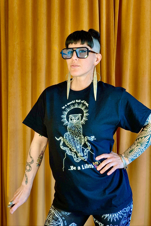 Model wearing a black "Be a Lilith Tee" with a bold owl and tentacle design, oversized glasses, large earrings, and tattooed arms against a mustard yellow curtain backdrop.