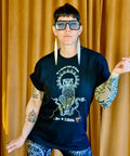 Person wearing a black "Be a Lilith Tee" featuring an intricate owl design and text, paired with black leggings covered in white mystical symbols, standing against a mustard curtain.