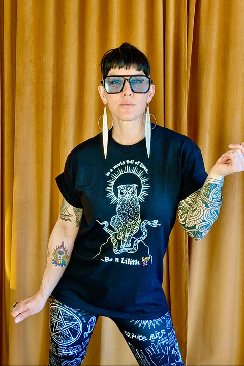 Person wearing a black "Be a Lilith Tee" featuring an intricate owl design and text, paired with black leggings covered in white mystical symbols, standing against a mustard curtain.