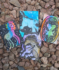 Four small illustrated cards or tags with colorful fantasy animal artwork are placed on a bed of small brown and gray pebbles outdoors.