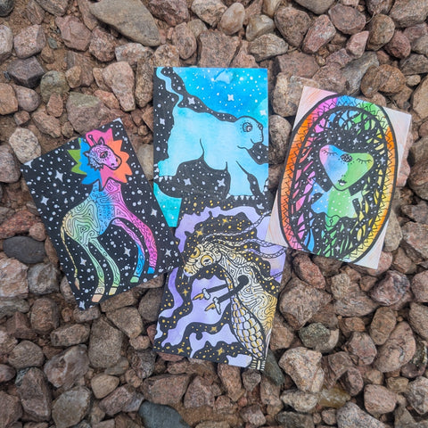 Four small illustrated cards or tags with colorful fantasy animal artwork are placed on a bed of small brown and gray pebbles outdoors.