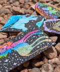 Four square cards with colorful psychedelic cosmic illustrations scattered on a bed of brown and gray pebbles.