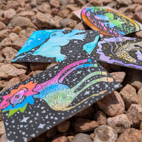 Four square cards with colorful psychedelic cosmic illustrations scattered on a bed of brown and gray pebbles.