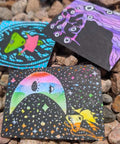 Three small square art cards or prints with colorful psychedelic illustrations, placed on a bed of brown and gray pebbles outdoors.
