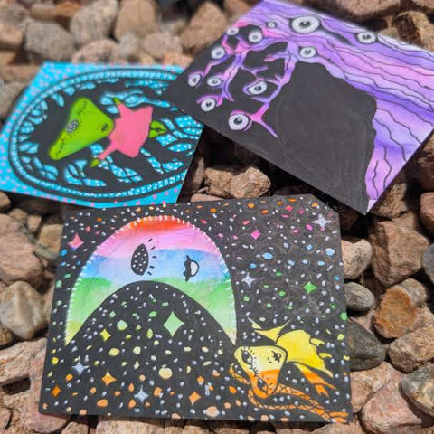 Three small square art cards or prints with colorful psychedelic illustrations, placed on a bed of brown and gray pebbles outdoors.