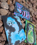 Three illustrated cards or art pieces with black backgrounds and colorful abstract creature designs, placed on a bed of brown pebbles outdoors.