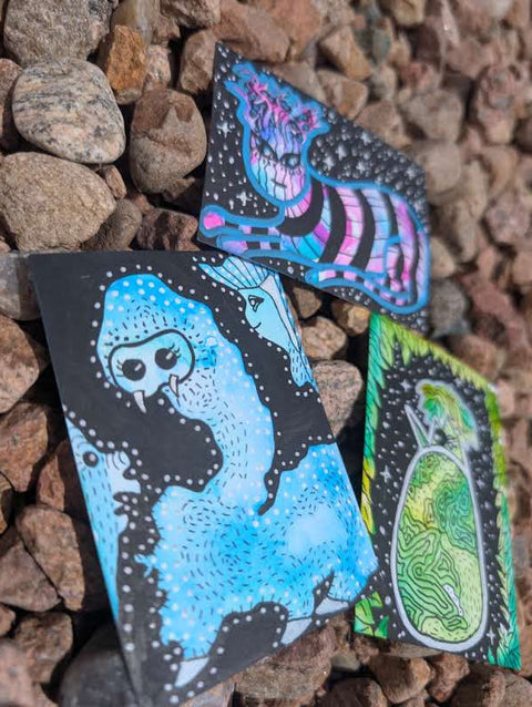 Three illustrated cards or art pieces with black backgrounds and colorful abstract creature designs, placed on a bed of brown pebbles outdoors.
