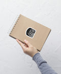 A hand holds a brown spiral notebook against a white textured wall, featuring a black and white As Above, So Below Sticker (B&W) with celestial and esoteric design printed on durable vinyl.