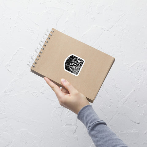 A hand holds a brown spiral notebook against a white textured wall, featuring a black and white As Above, So Below Sticker (B&W) with celestial and esoteric design printed on durable vinyl.