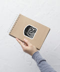 Hand holding a brown spiral notebook with a black and white "As Above, So Below Sticker (B&W)" featuring mystical symbols, printed on durable adhesive vinyl.