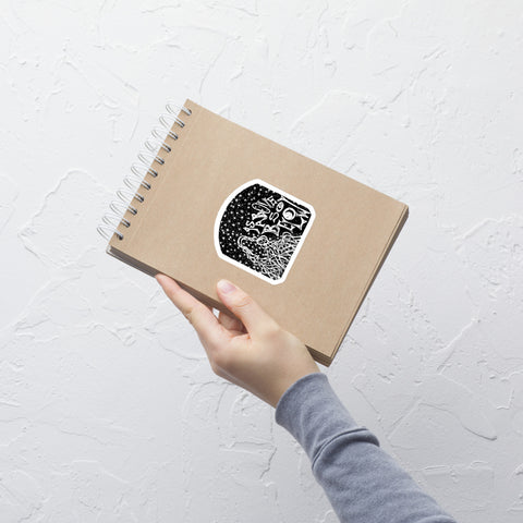 Hand holding a brown spiral notebook with a black and white "As Above, So Below Sticker (B&W)" featuring mystical symbols, printed on durable adhesive vinyl.