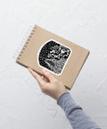 Hand holding a spiral-bound kraft notebook with an "As Above, So Below Sticker (B&W)" featuring intricate celestial and symbolic black and white artwork on the cover against a textured white wall.