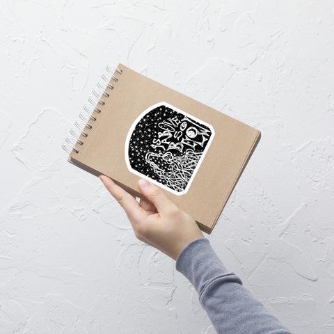 Hand holding a spiral-bound kraft notebook with an "As Above, So Below Sticker (B&W)" featuring intricate celestial and symbolic black and white artwork on the cover against a textured white wall.