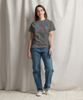 A person wearing an "All Eyes On Me Tee" in solid charcoal color featuring a colorful eye and leaf pattern on the front, paired with blue jeans and white sneakers, standing against a neutral curtain backdrop.