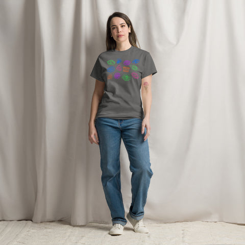 A person wearing an "All Eyes On Me Tee" in solid charcoal color featuring a colorful eye and leaf pattern on the front, paired with blue jeans and white sneakers, standing against a neutral curtain backdrop.