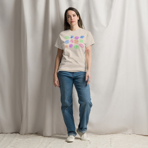 Woman wearing a neutral-colored All Eyes On Me Tee featuring colorful eye and lip illustrations, paired with blue jeans and white sneakers, standing against a soft draped background.